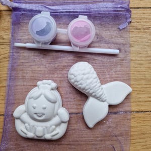 Puede incluir: Una bolsa de malla morada llena de dos peque&ntilde;os botes de pintura, un pincel y dos figuritas de cer&aacute;mica blanca. Una figurita es una cola de sirena y la otra es una ni&ntilde;a con una corona.