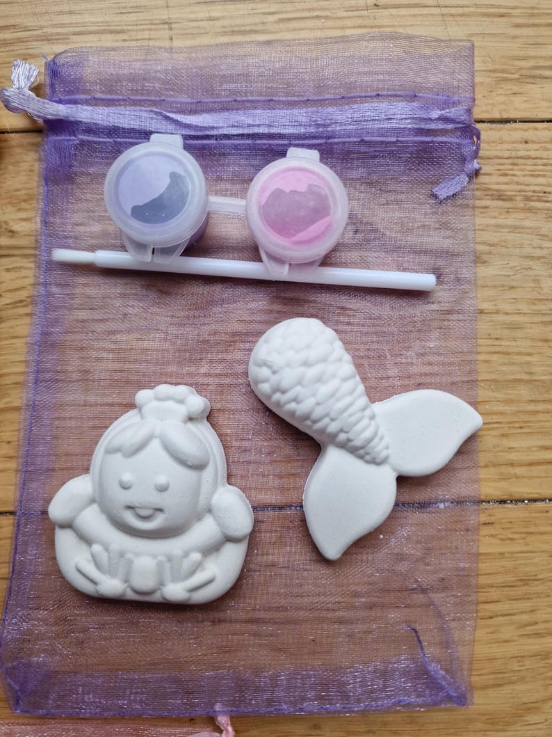 Puede incluir: Una bolsa de malla morada llena de dos peque&ntilde;os botes de pintura, un pincel y dos figuritas de cer&aacute;mica blanca. Una figurita es una cola de sirena y la otra es una ni&ntilde;a con una corona.