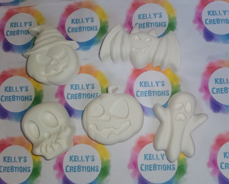 May include: Set of six white ceramic Halloween-themed ornaments. The ornaments include a pumpkin, a ghost, a bat, a skull, and two more pumpkins. Each ornament has a blank surface for painting or decorating.