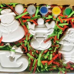 May include: A Christmas-themed paint-your-own kit in a cardboard box. The kit includes unpainted ceramic figures of a snowman, reindeer, Santa Claus, and a sleigh, along with paint pots and a paintbrush. The figures are nestled in red and green shredded paper.