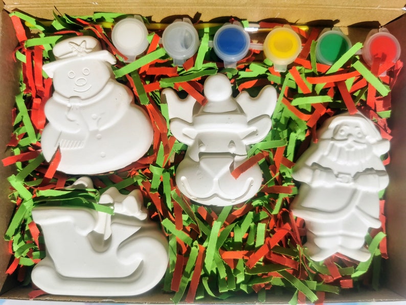 May include: A Christmas-themed paint-your-own kit in a cardboard box. The kit includes unpainted ceramic figures of a snowman, reindeer, Santa Claus, and a sleigh, along with paint pots and a paintbrush. The figures are nestled in red and green shredded paper.