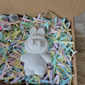 Peut inclure: Une figurine de lapin en c&eacute;ramique blanche avec un visage souriant, &agrave; l'int&eacute;rieur d'une bo&icirc;te en carton remplie de papier d&eacute;chiquet&eacute; color&eacute;. Le lapin a de grandes oreilles et est pr&ecirc;t &agrave; &ecirc;tre peint. La bo&icirc;te contient &eacute;galement de petits pots de peinture.