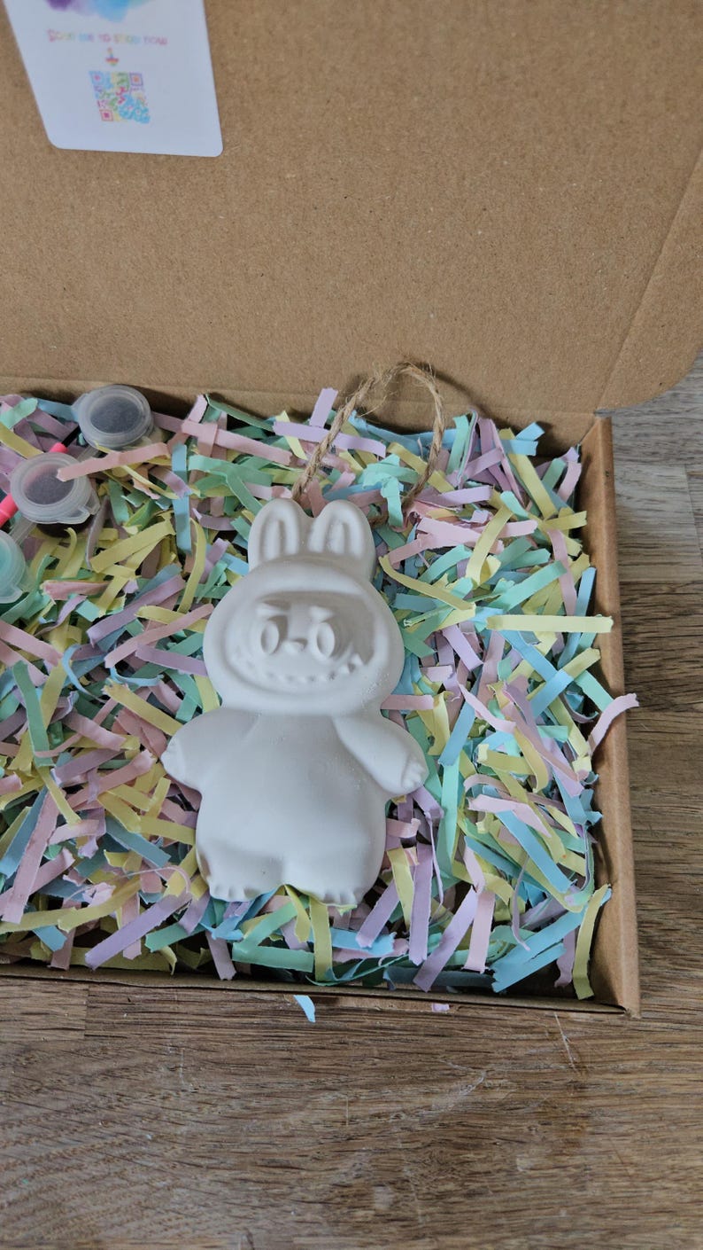Peut inclure: Une figurine de lapin en c&eacute;ramique blanche avec un visage souriant, &agrave; l'int&eacute;rieur d'une bo&icirc;te en carton remplie de papier d&eacute;chiquet&eacute; color&eacute;. Le lapin a de grandes oreilles et est pr&ecirc;t &agrave; &ecirc;tre peint. La bo&icirc;te contient &eacute;galement de petits pots de peinture.