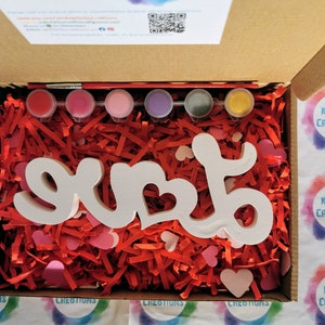May include: A wooden "LOVE" sign with a heart cutout, ready to be painted. The sign is surrounded by red and pink shredded paper and comes with six paint pots and a paintbrush.