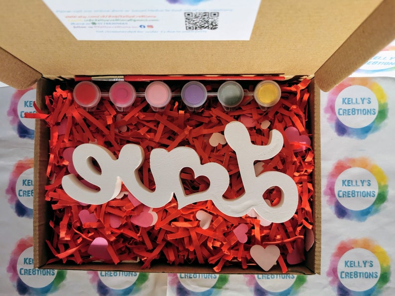 May include: A wooden "LOVE" sign with a heart cutout, ready to be painted. The sign is surrounded by red and pink shredded paper and comes with six paint pots and a paintbrush.