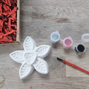 May include: A white, five-petaled flower-shaped plaster figure with leaf-like textures, accompanied by three small plastic paint pots containing white, pink, and black paint, and a thin paintbrush with a red handle and black bristles.