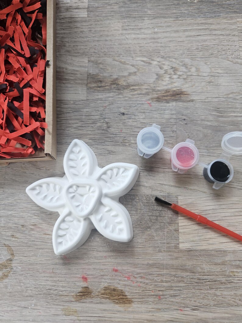 May include: A white, five-petaled flower-shaped plaster figure with leaf-like textures, accompanied by three small plastic paint pots containing white, pink, and black paint, and a thin paintbrush with a red handle and black bristles.