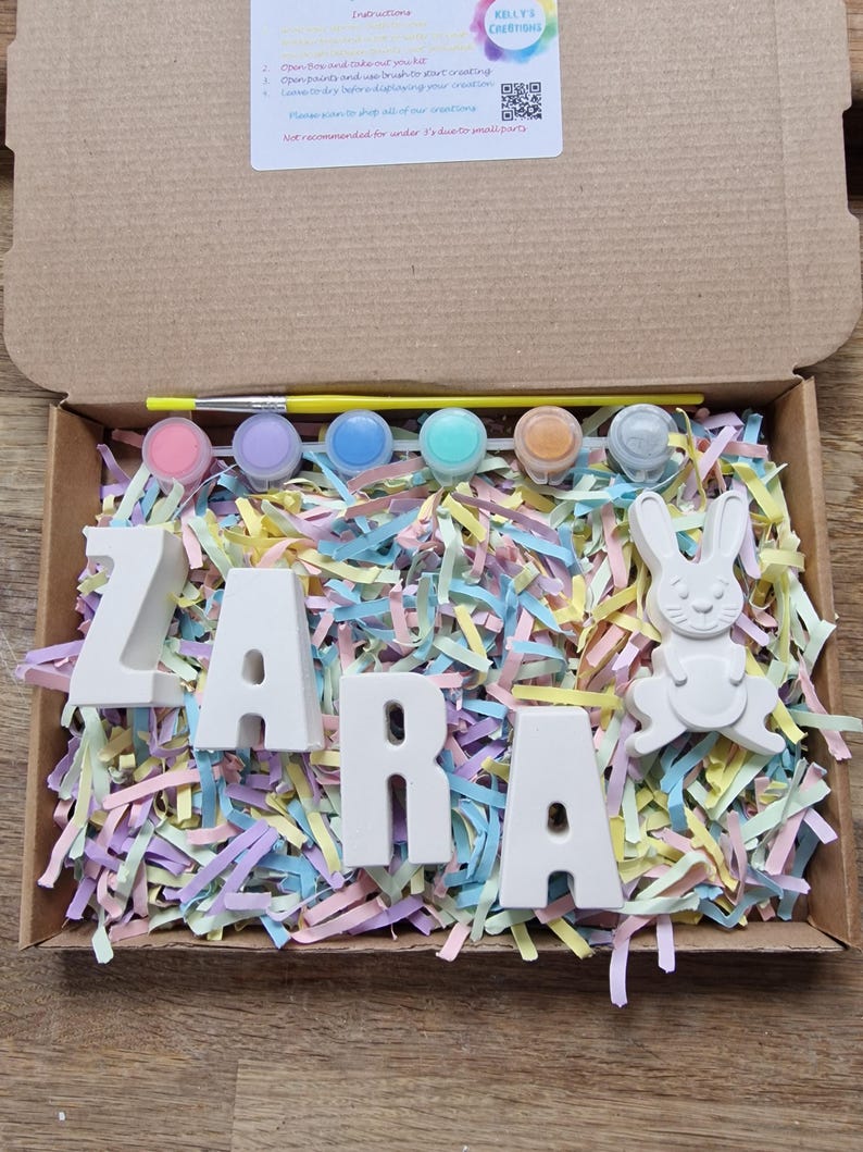 May include: A craft kit for painting ceramic letters that spell out the name "ZARA" with a small ceramic bunny figurine. The kit includes six small paint pots, a paintbrush, and colorful shredded paper.