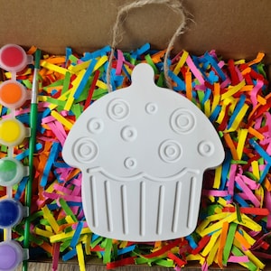 May include: A white ceramic cupcake-shaped ornament with a string for hanging. The ornament has a raised design of circles and is ready to be painted. The ornament is surrounded by colourful paper shreds.