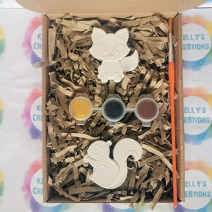 May include: A craft kit with two white ceramic animal figurines, a fox and a squirrel, ready to be painted. The kit includes three small paint pots, one yellow, one black, and one brown, and a paintbrush.