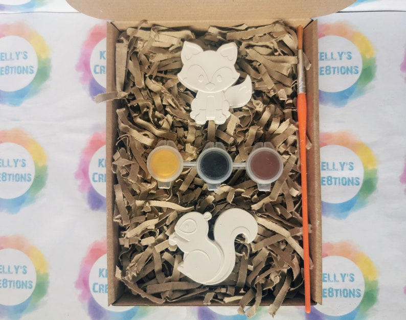 May include: A craft kit with two white ceramic animal figurines, a fox and a squirrel, ready to be painted. The kit includes three small paint pots, one yellow, one black, and one brown, and a paintbrush.