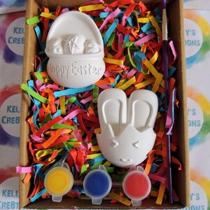 May include: A craft kit featuring two white ceramic Easter-themed figurines, a basket and a bunny, with paint pots in yellow, blue, and red, and a paintbrush. The figurines are ready to be painted.