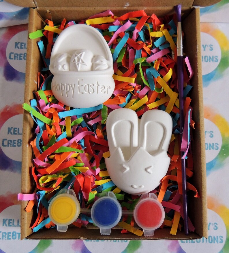 May include: A craft kit featuring two white ceramic Easter-themed figurines, a basket and a bunny, with paint pots in yellow, blue, and red, and a paintbrush. The figurines are ready to be painted.