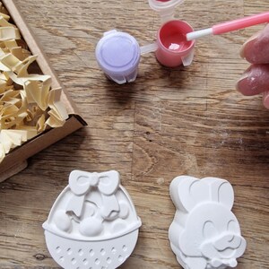 May include: Two white ceramic Easter-themed figurines, one shaped like a basket with a bow and eggs, the other shaped like a bunny with a bow tie.  Two small paint pots, one purple and one pink, with paintbrushes.