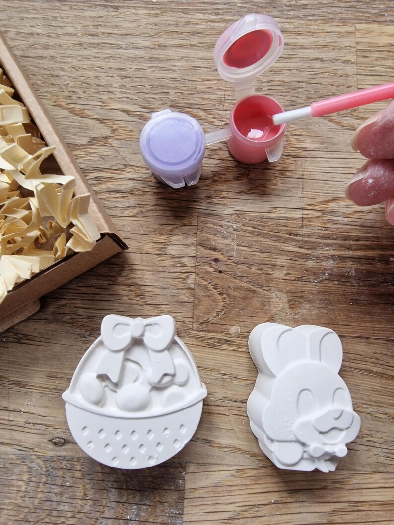 May include: Two white ceramic Easter-themed figurines, one shaped like a basket with a bow and eggs, the other shaped like a bunny with a bow tie.  Two small paint pots, one purple and one pink, with paintbrushes.