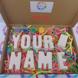 May include: A craft kit in a cardboard box, featuring the words "YOUR NAME" in white block letters, paint pots, and a white building block. The box is filled with colorful shredded paper. The text "Set: N2" is visible.