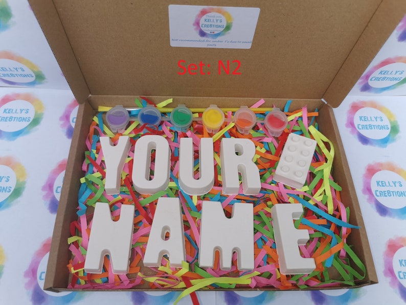 May include: A craft kit in a cardboard box, featuring the words "YOUR NAME" in white block letters, paint pots, and a white building block. The box is filled with colorful shredded paper. The text "Set: N2" is visible.