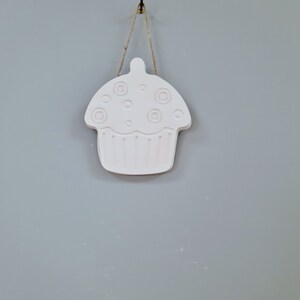 May include: A white ceramic cupcake ornament with a string hanging from a hook. The cupcake has a swirl design on top and a scalloped edge.