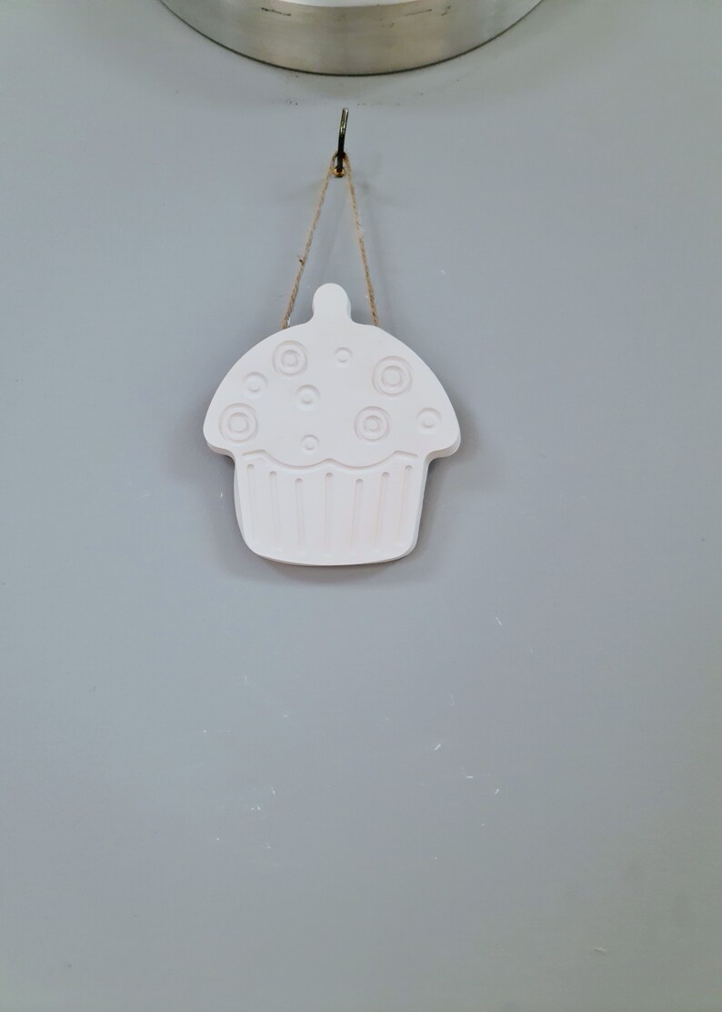 May include: A white ceramic cupcake ornament with a string hanging from a hook. The cupcake has a swirl design on top and a scalloped edge.