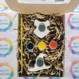 May include: A craft kit in a cardboard box, containing two white ceramic shapes, paint pots in black, yellow, and red, and a yellow paintbrush. The box is filled with brown paper shreds. The text "KELLY'S CREATIONS" is visible.