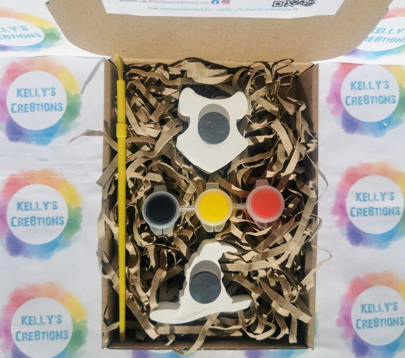 May include: A craft kit in a cardboard box, containing two white ceramic shapes, paint pots in black, yellow, and red, and a yellow paintbrush. The box is filled with brown paper shreds. The text "KELLY'S CREATIONS" is visible.