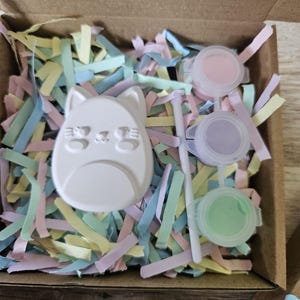 Puede incluir: Un kit de manualidades en una caja de cart&oacute;n que contiene una figura de gato de yeso blanco, un pincel blanco peque&ntilde;o y tres botes de pintura de pl&aacute;stico redondos con pintura rosa, morada y verde claro, sobre tiras de papel de colores pastel.