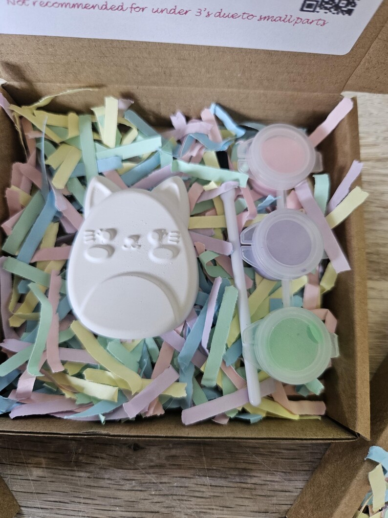 Puede incluir: Un kit de manualidades en una caja de cart&oacute;n que contiene una figura de gato de yeso blanco, un pincel blanco peque&ntilde;o y tres botes de pintura de pl&aacute;stico redondos con pintura rosa, morada y verde claro, sobre tiras de papel de colores pastel.