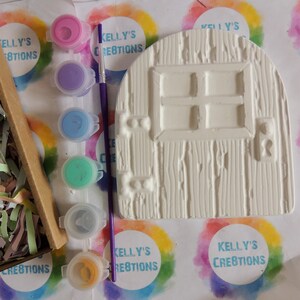 May include: A white ceramic door with a window and a keyhole, along with five small paint pots in purple, blue, light blue, green, and yellow, and a purple paintbrush. The paint pots and brush are on a white surface with colorful rainbow circles.
