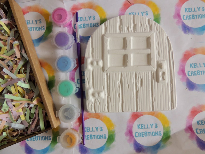May include: A white ceramic door with a window and a keyhole, along with five small paint pots in purple, blue, light blue, green, and yellow, and a purple paintbrush. The paint pots and brush are on a white surface with colorful rainbow circles.