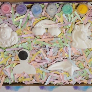Puede incluir: Una caja llena de virutas de papel de colores y figuritas de cer&aacute;mica blanca, incluyendo un unicornio, un arco&iacute;ris, una estrella, un mu&ntilde;eco de nieve y un astronauta. La caja tambi&eacute;n incluye peque&ntilde;os recipientes de pintura.