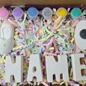 May include: A craft kit for painting ceramic Easter decorations. The kit includes a bunny, an egg, and the letters N-A-M-E. The kit also includes six small pots of paint in pink, purple, blue, gray, yellow, and green, and a paintbrush.