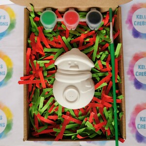 May include: A white ceramic snowman ornament with a paintbrush, red, green, and black paint pots, and green and red shredded paper in a brown box. The box is labeled "Kelly's Creations".