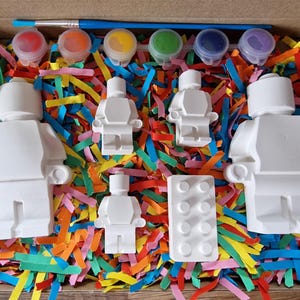 May include: A set of five white plastic figurines of people, a white plastic brick, and six small paint pots with lids. The paint pots are red, blue, green, yellow, purple, and orange. The figurines and brick are sitting on a bed of colorful shredded paper.