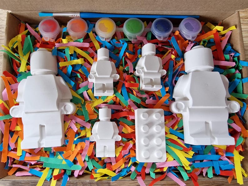 May include: A set of five white plastic figurines of people, a white plastic brick, and six small paint pots with lids. The paint pots are red, blue, green, yellow, purple, and orange. The figurines and brick are sitting on a bed of colorful shredded paper.