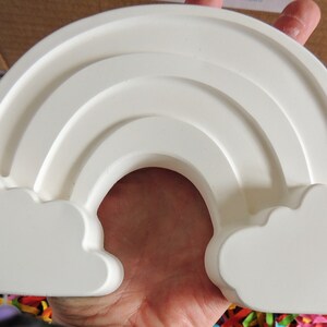 May include: White ceramic rainbow with two clouds on either side. The rainbow has three arches and is a popular craft project.
