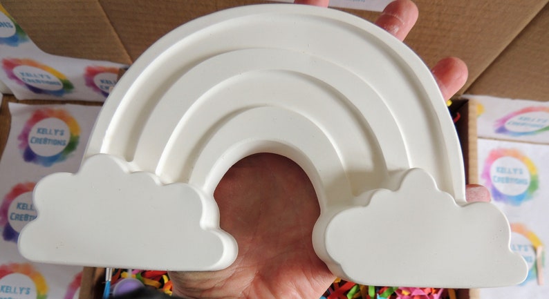 May include: White ceramic rainbow with two clouds on either side. The rainbow has three arches and is a popular craft project.