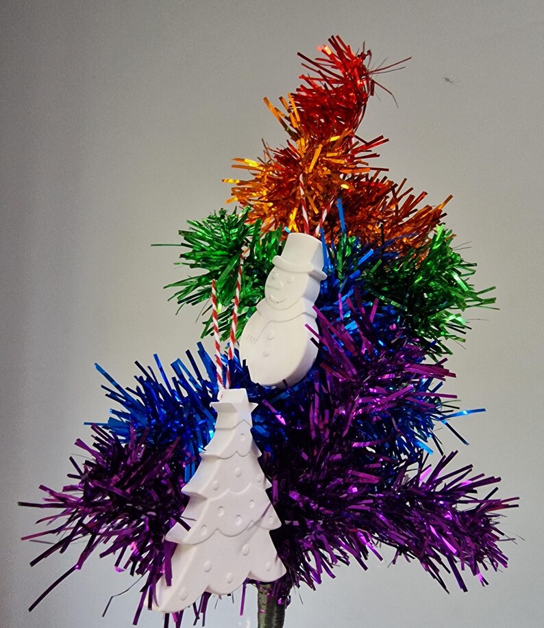 May include: White ceramic Christmas ornaments in the shape of a snowman and a Christmas tree hanging from a rainbow tinsel tree.
