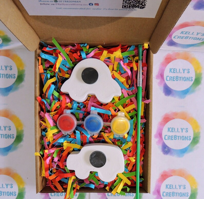 Puede incluir: Un kit de manualidades en una caja de cart&oacute;n, con dos imanes blancos con forma de coche, tres botes de pintura en rojo, azul y amarillo, y un pincel verde. La caja est&aacute; llena de papel triturado de colores.