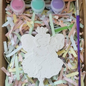 May include: A white ceramic fairy figurine with wings, a crown, and a dress. The figurine is in a box with pink, green, and purple paint pots and a purple paintbrush. The box is filled with colorful shredded paper.