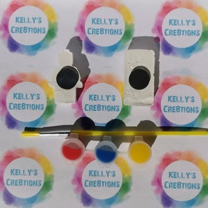Puede incluir: Un kit de pintura por n&uacute;meros con dos figuritas de cer&aacute;mica, una de un mu&ntilde;eco de nieve y otra de una casa. El kit incluye cuatro peque&ntilde;os botes de pintura, un pincel y ocho pegatinas circulares con el texto "KELLY'S CREATIONS" impreso en ellas.