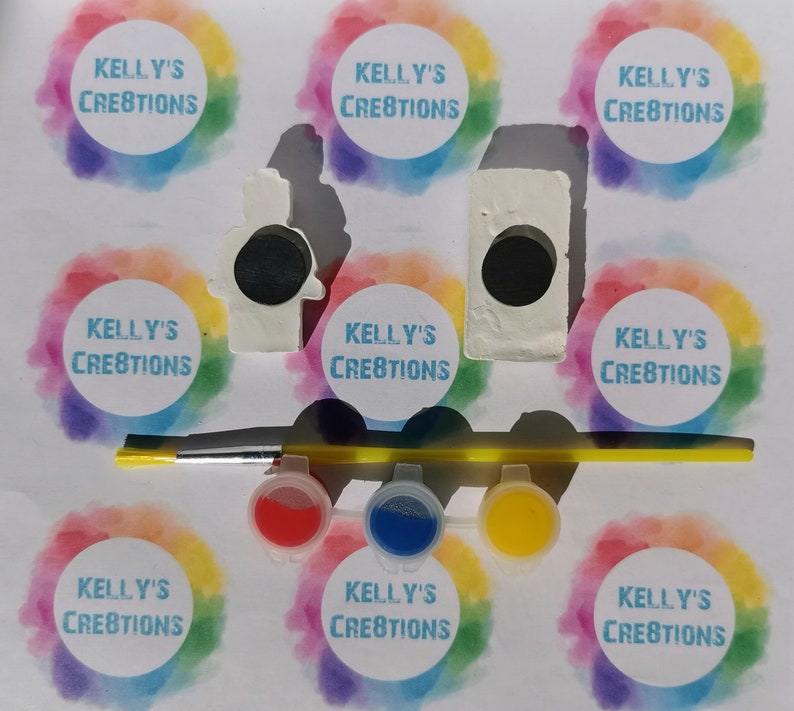 Puede incluir: Un kit de pintura por n&uacute;meros con dos figuritas de cer&aacute;mica, una de un mu&ntilde;eco de nieve y otra de una casa. El kit incluye cuatro peque&ntilde;os botes de pintura, un pincel y ocho pegatinas circulares con el texto "KELLY'S CREATIONS" impreso en ellas.