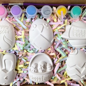 May include: A set of six white ceramic Easter egg ornaments with different designs, including a bunny, a basket of eggs, and a banner that says "Happy Easter". The ornaments are in a box with colourful shredded paper and six small paint pots with different colours.