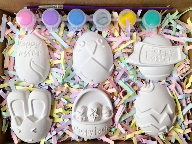 May include: A set of six white ceramic Easter egg ornaments with different designs, including a bunny, a basket of eggs, and a banner that says "Happy Easter". The ornaments are in a box with colourful shredded paper and six small paint pots with different colours.