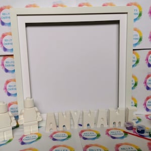 May include: A white picture frame with the word "ANYNAME" in individual letters. Two white Lego-style figures stand near the frame. Small paint pots and a paintbrush are also present, suggesting a DIY craft kit.