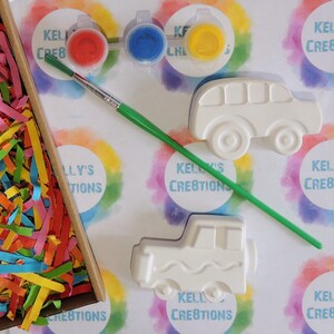 Puede incluir: Un kit de manualidades con figuras de cer&aacute;mica con forma de autob&uacute;s y cami&oacute;n, botes de pintura roja, azul y amarilla y un pincel. El kit incluye papel triturado de colores y el texto "Kelly's Creations".