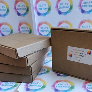 May include: A stack of four brown cardboard boxes with a single box on the right. The box on the right has a sticker that says "Thank you for coming to my party! Ruby". The boxes are on a white surface with colorful circles that say "Kelly's Creations".