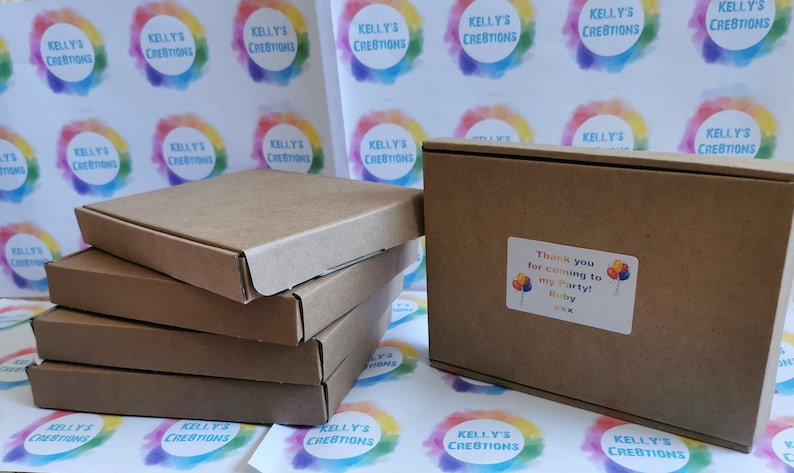 May include: A stack of four brown cardboard boxes with a single box on the right. The box on the right has a sticker that says "Thank you for coming to my party! Ruby". The boxes are on a white surface with colorful circles that say "Kelly's Creations".