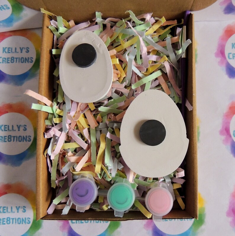 May include: A craft kit containing two white egg-shaped wooden ornaments, each with a black circular magnet. The kit includes three small paint pots in purple, green, and pink. The egg ornaments are nestled in pastel-coloured shredded paper inside a cardboard box.