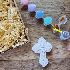 May include: A white ceramic cross with a Celtic knot design, ready to be painted with the included set of colorful acrylic paints. The paints come in a variety of colors, including pink, purple, blue, turquoise, gold, and gray.