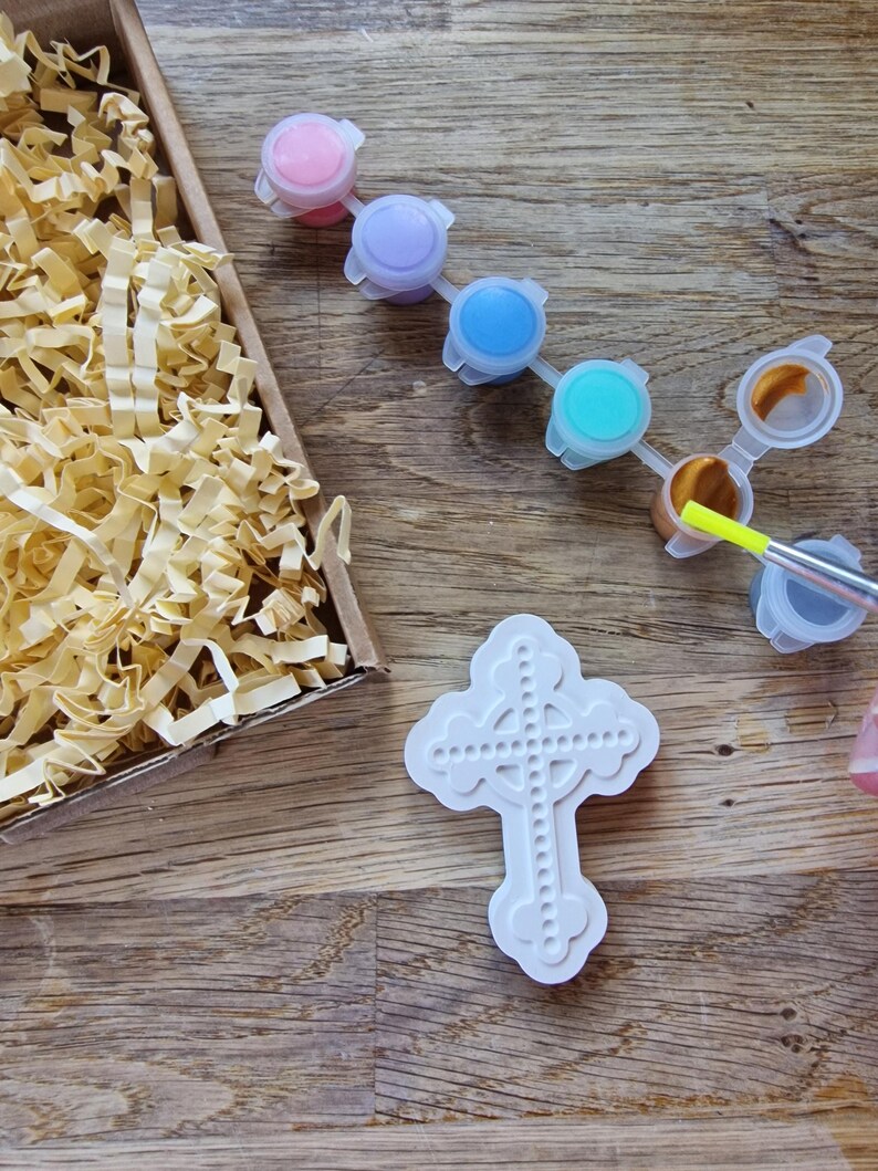 May include: A white ceramic cross with a Celtic knot design, ready to be painted with the included set of colorful acrylic paints. The paints come in a variety of colors, including pink, purple, blue, turquoise, gold, and gray.